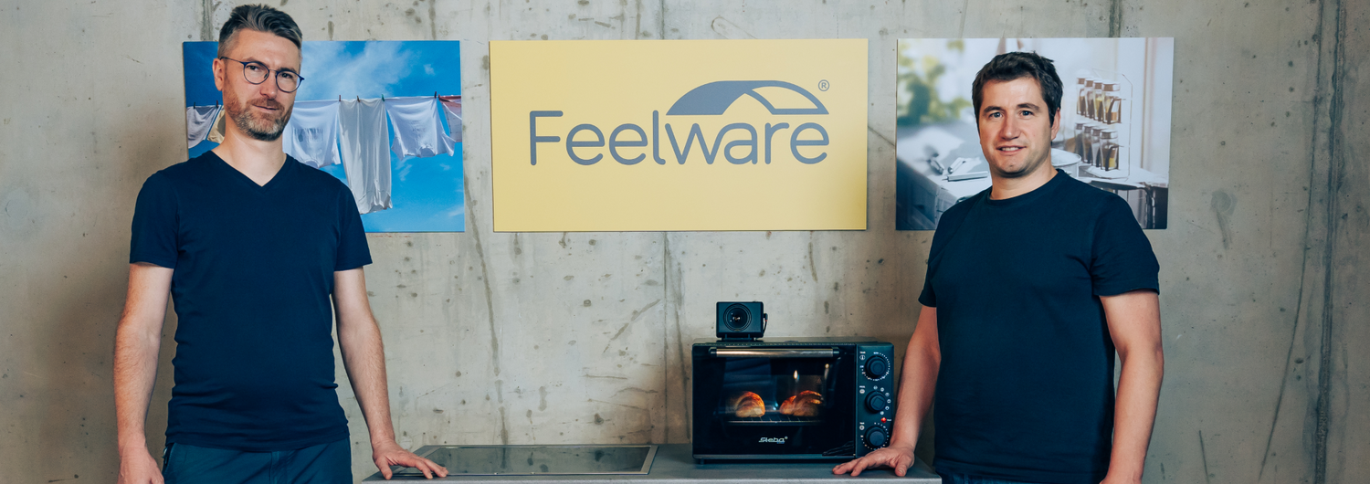 The founders of Feelware, Jean and Robert, stand in the picture. Between themDie Gründer von Feelware, Jean und Robert, stehen auf dem Bild. Zwischen ihnen stehen ein Einbauherd und ein Mini-Ofen. Das Firmenlogo von Feelware ist an der Wand im Hintergrun