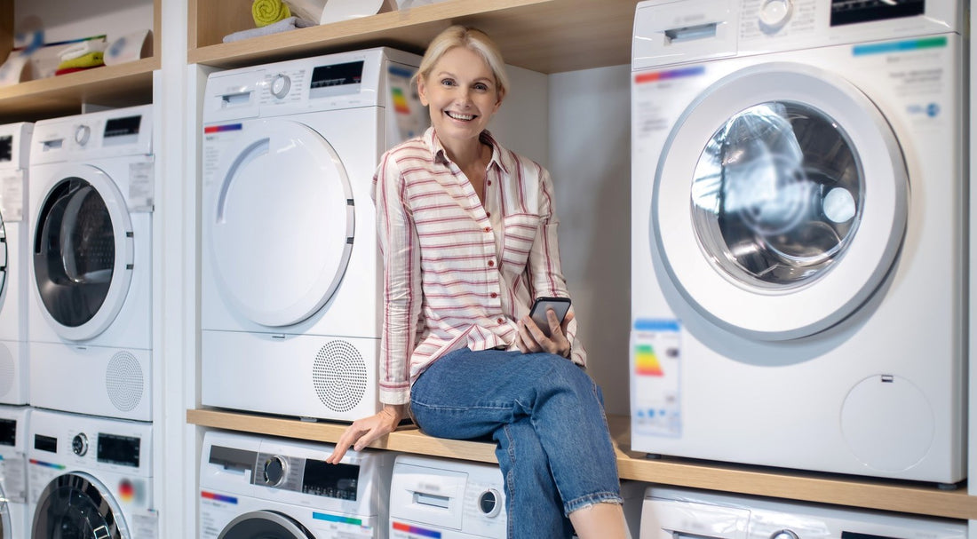 5 Waschmaschinen stehen gestapelt in einem Regal. Auf einer weiteren Waschmaschine sitzt eine lächelnde Frau.